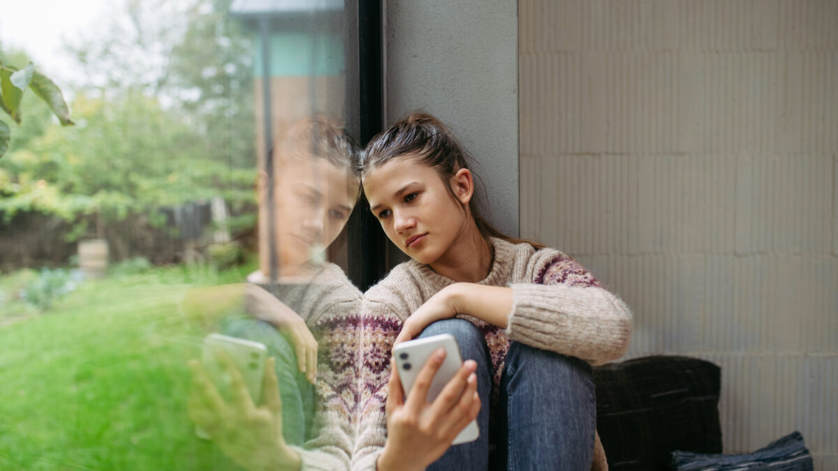 Teenage girl sitting by window holding smartphone in hand.