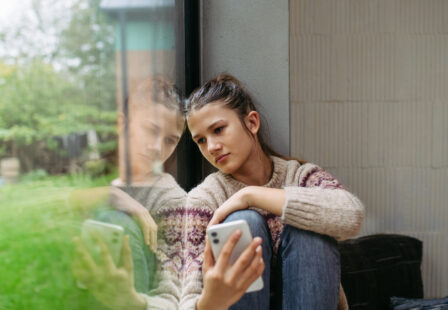 Teenage girl sitting by window holding smartphone in hand.