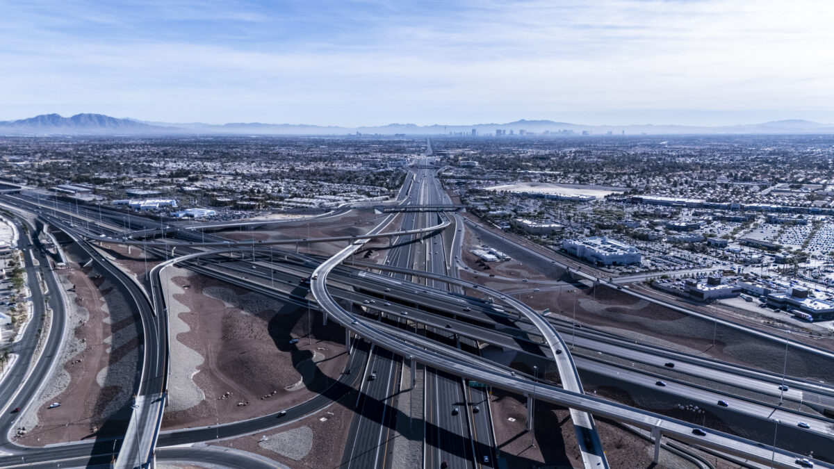 An elevated aerial image capturing the expansive freeway system leading into Las Vegas. The intricate network of roads and overpasses integrates seamlessly into the desert surroundings, with the distant city skyline visible on the horizon.
