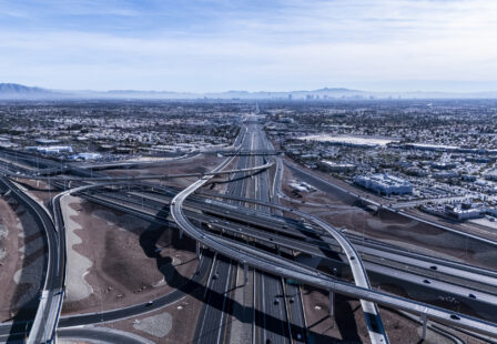 An elevated aerial image capturing the expansive freeway system leading into Las Vegas. The intricate network of roads and overpasses integrates seamlessly into the desert surroundings, with the distant city skyline visible on the horizon.